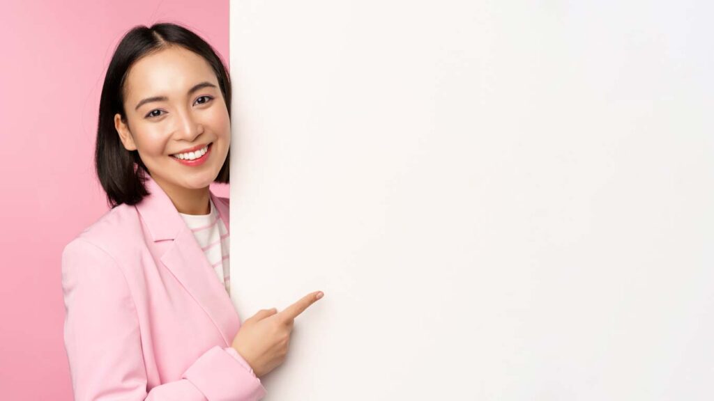 Image of asian female entrepreneur in suit, pointing finger at board, showing smth on white wall, demonstrating chart or information, standing over pink studio background.