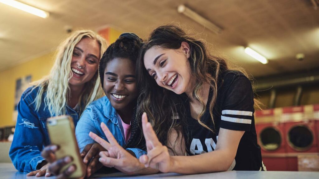 Smiling young female micro influencers leaning on a laundromat counter taking selfies while doing laundry together