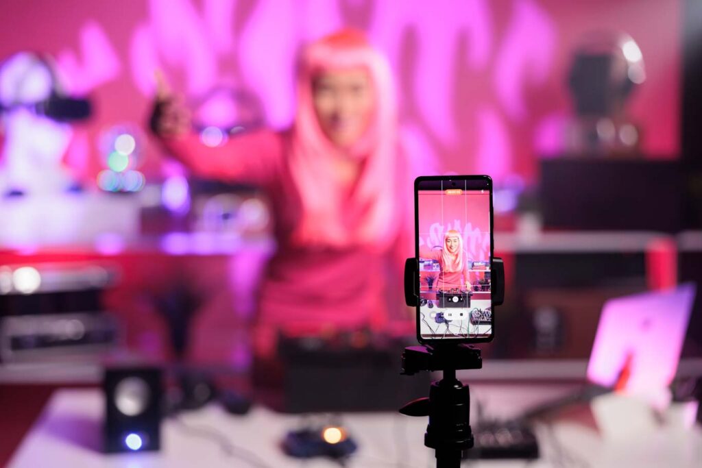 female micro influencer with pink hair playing techno song at professional turntables while filming music process with phone camera, having fun in studio. Dj artist doing performance at nightclub with audio equipment